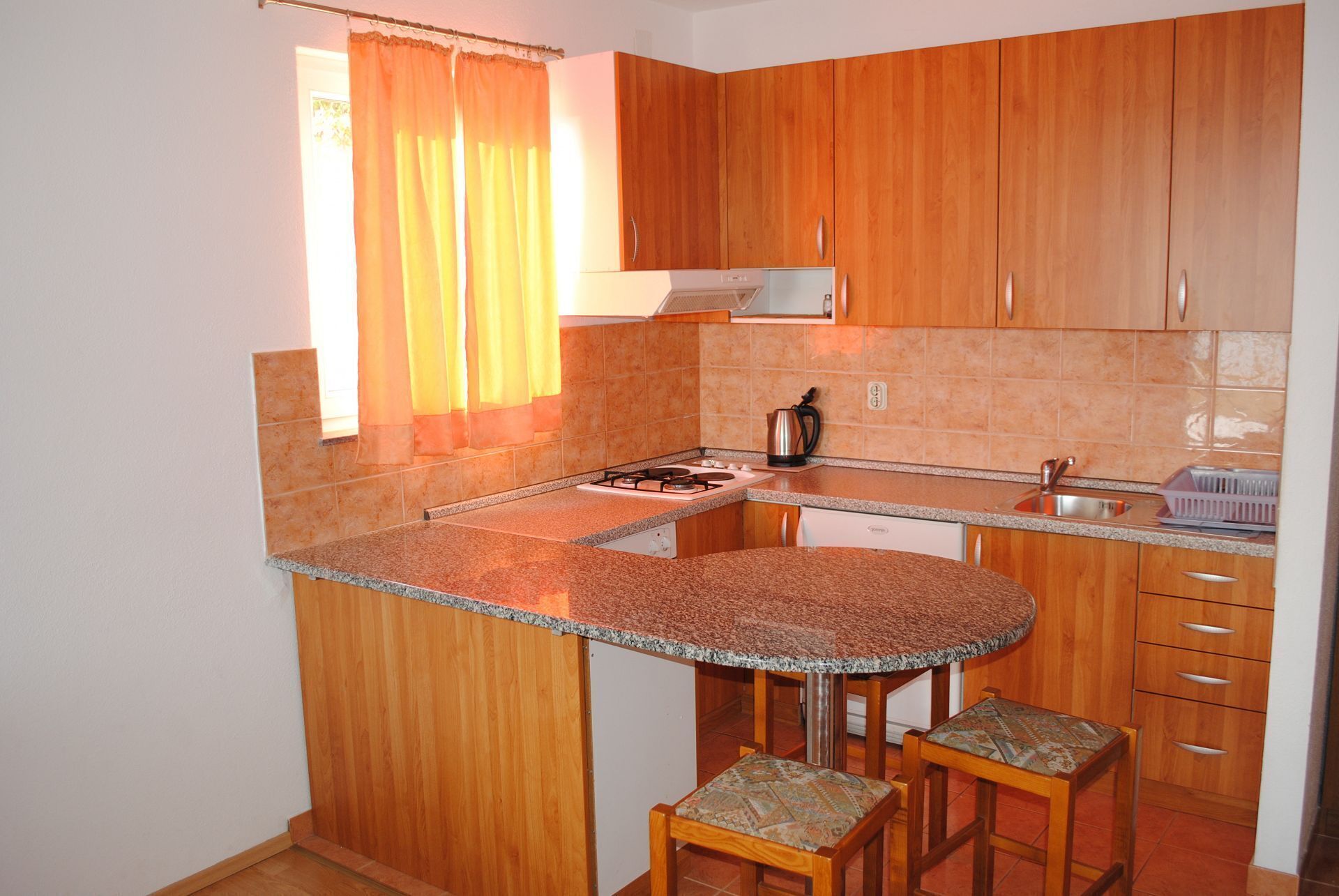 A compact kitchen with wooden cabinets, a granite countertop, a gas stove, a kettle, and two bar stools.