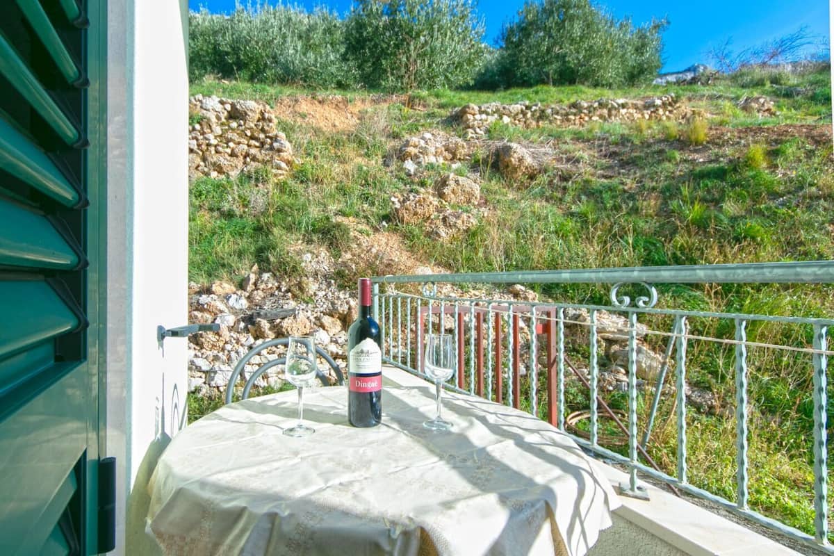 A balcony with a table set for two, a bottle of wine, and a glass. The balcony overlooks a rocky hillside with vegetation.