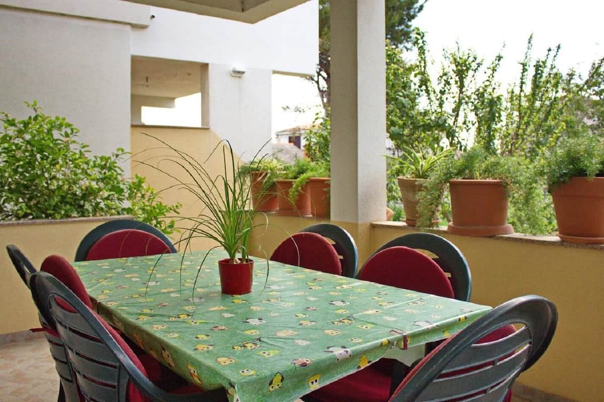 An outdoor patio with a table covered in a green tablecloth with cartoon characters, surrounded by red chairs. There are potted plants on the table and along the wall.