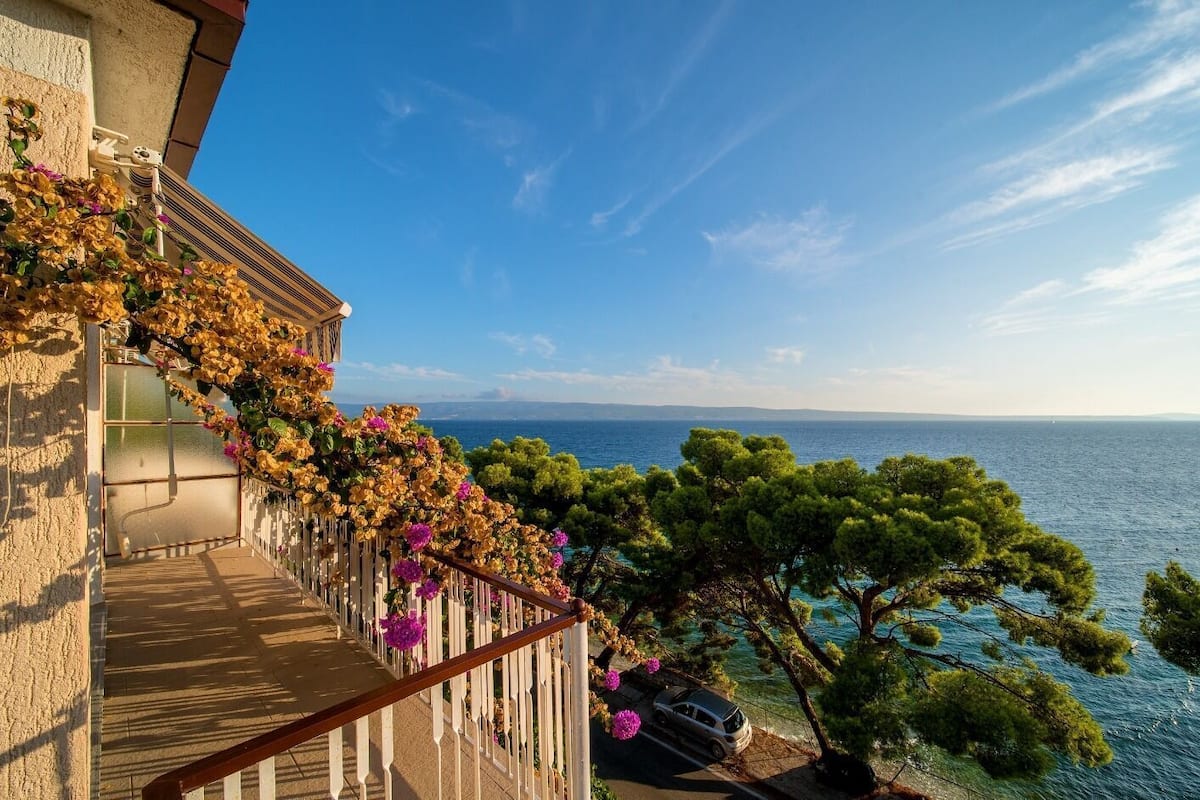 A balcony with a view of the sea, a car parked nearby, and a building with a textured facade.