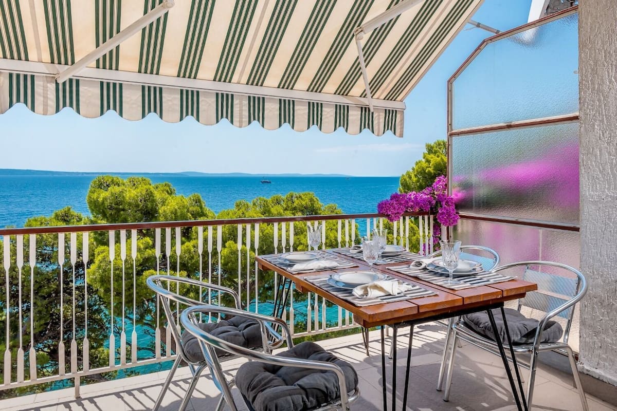 A balcony with a table set for dining, overlooking a scenic view of the sea and greenery.