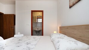 A bedroom with a wooden headboard, a bed with a marble-patterned bedspread, a nightstand with a lamp, and a bathroom visible through an open door. - Apartments Ante (Seget)