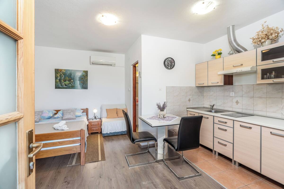 A compact room with a bed, a small dining table, a kitchen area with cabinets and a sink, and a painting on the wall.