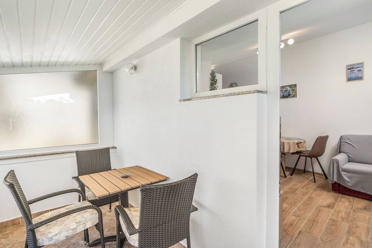A room with a wooden table and wicker chairs, a grey sofa, and a small window with a view.