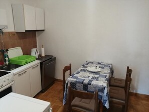 Uma cozinha compacta com armários brancos, um escorredor de louça verde, uma cafeteira e uma mesa com toalha de mesa azul e branca.