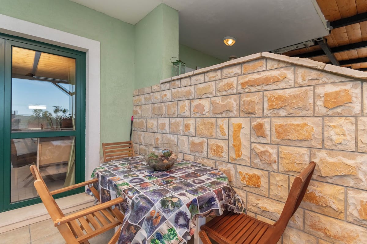 A dining area with a table covered by a patterned tablecloth, surrounded by wooden chairs, against a stone wall.