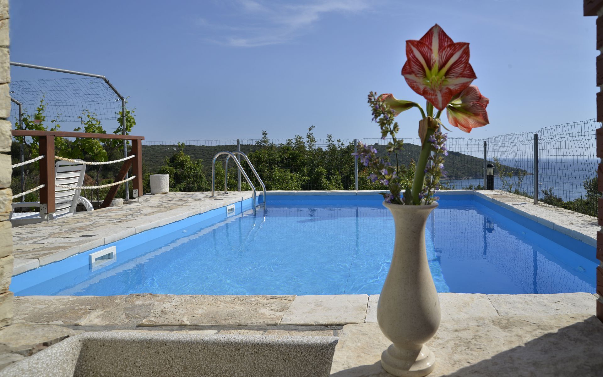 A clear blue swimming pool with a stone deck, a white vase with flowers, and a view of the sea and hills in the background.