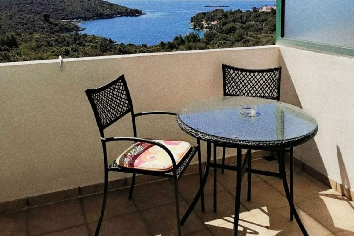 A balcony with a round glass table and two chairs, overlooking a coastal landscape with a clear view of the sea and hills.