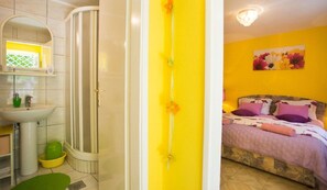 A bathroom with a shower, sink, and toilet. There is a bed with a headboard and pillows in the adjacent room. - Apartment Zelja (Orebic)