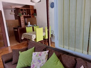 A living room with a brown sofa, green cushions, and a floral pillow. There is a dining area with a table and chairs, and a kitchen area with cabinets and a hanging light fixture. - Apartment Silvana (Sali)