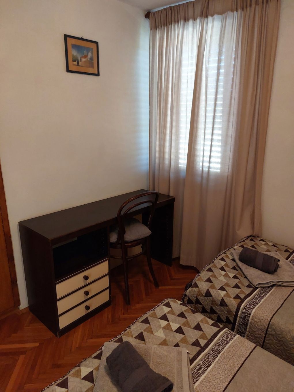 A bedroom with a wooden desk, a chair, a bed with patterned bedding, and a window with curtains.