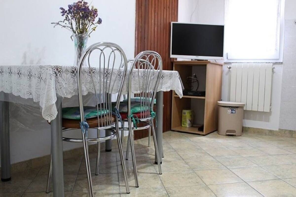 A dining area with a table covered by a lace tablecloth, two metal chairs with cushions, a vase with flowers, a television on a wooden stand, and a radiator on the wall.