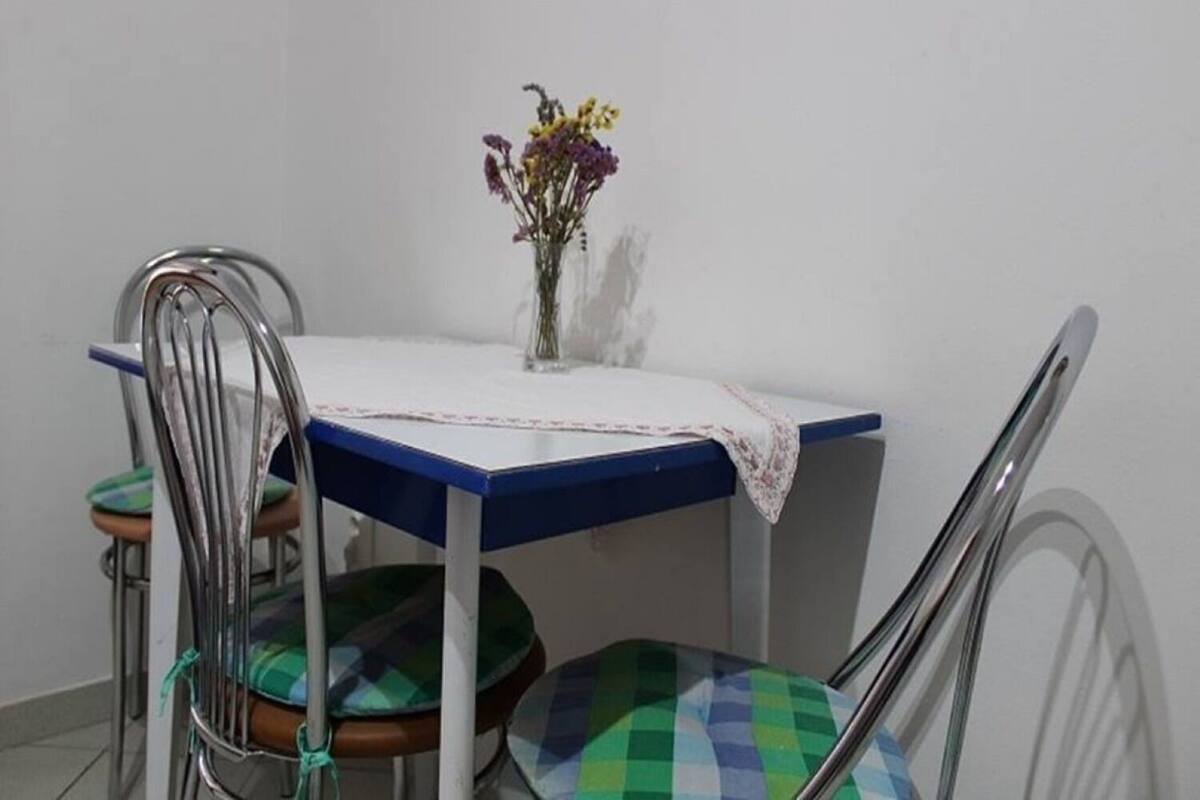 A dining table with a blue top and white tablecloth, surrounded by metal chairs with green and blue checkered cushions. A vase with flowers is placed on the table.