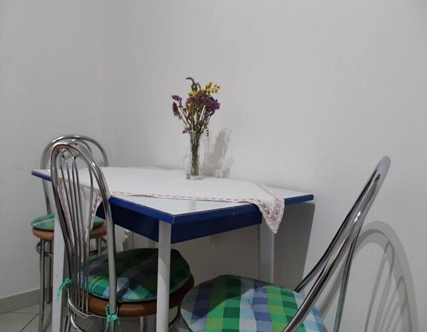 Ein Esstisch mit blauer Tischplatte und weißer Tischdecke, umgeben von Metallstühlen mit grün-blau karierten Kissen. Auf dem Tisch steht eine Vase mit Blumen.