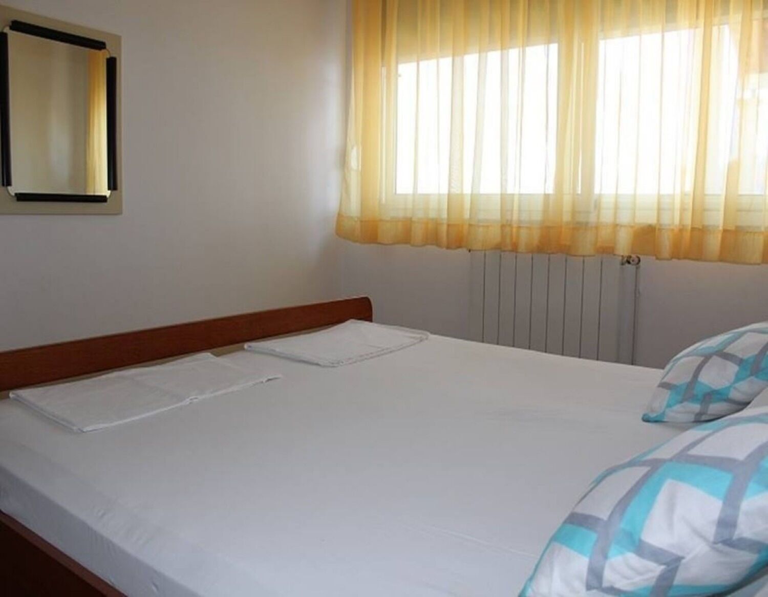 Una cama individual con cabecera de madera, una ventana con cortinas amarillas y un radiador debajo.