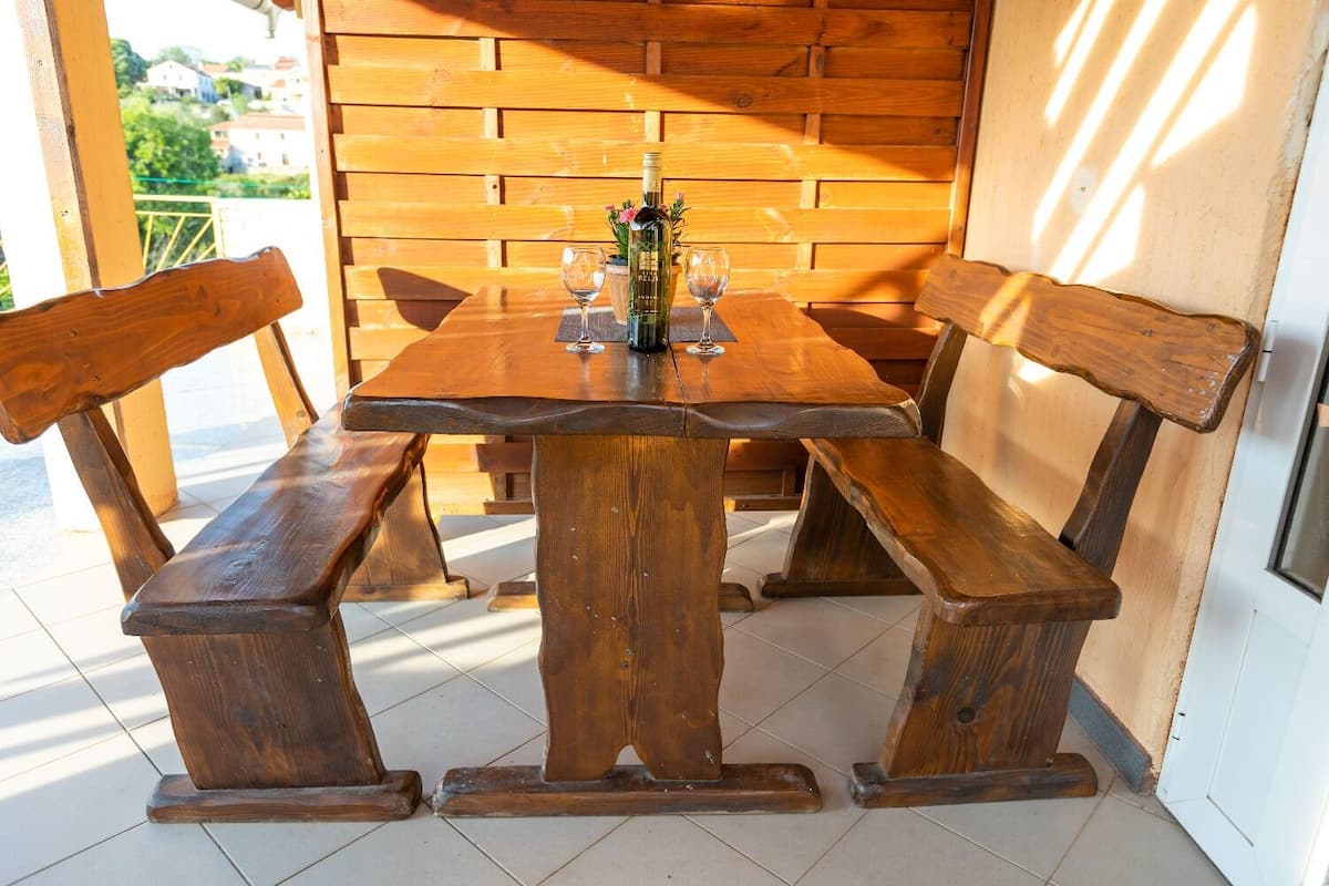 Una mesa de madera con bancos, una botella de vino y dos copas de vino en un patio con un muro de madera al fondo.