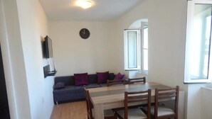 A compact living space with a dining table and chairs, a sofa with cushions, a clock on the wall, and a window providing natural light. - Apartment Antonio (Murter-Kornati)