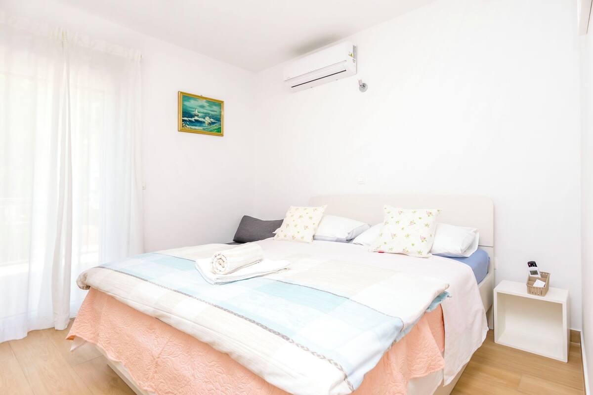 Un lit bien fait, recouvert d’une couvre-lit à rayures, une table de chevet avec une lampe et un climatiseur fixé au mur.