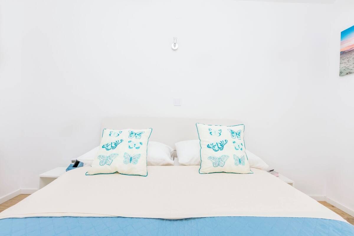 Un lit avec une literie blanche et des oreillers bleus, une table de chevet blanche et un luminaire fixé au mur.