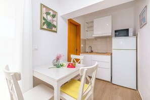 Cocina pequeña con armarios blancos, un microondas y un refrigerador. Hay una mesa de comedor con sillas y un florero con flores.