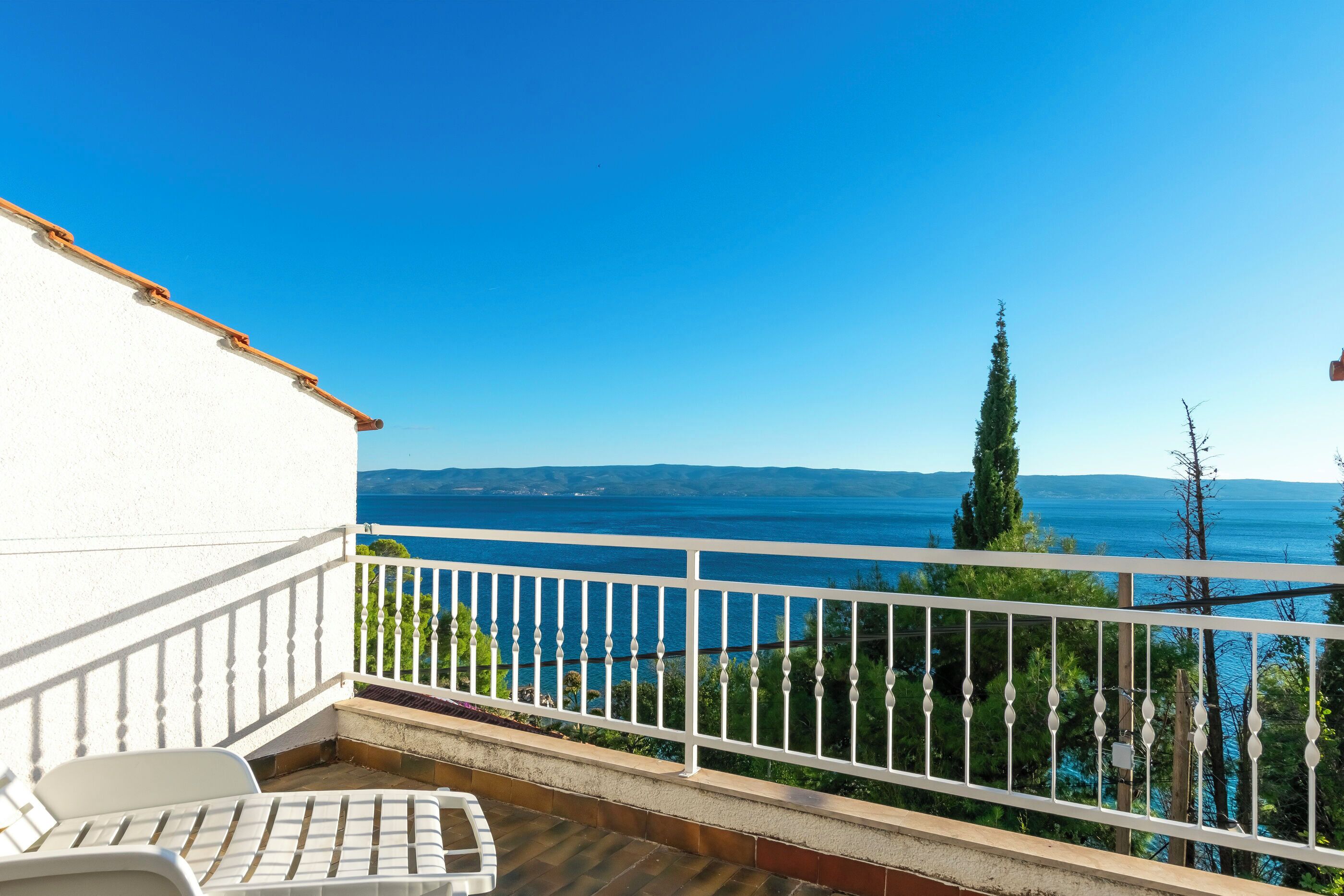 Ein Balkon mit weißer Reling, einem weißen Stuhl und Blick auf das Meer und die Berge.