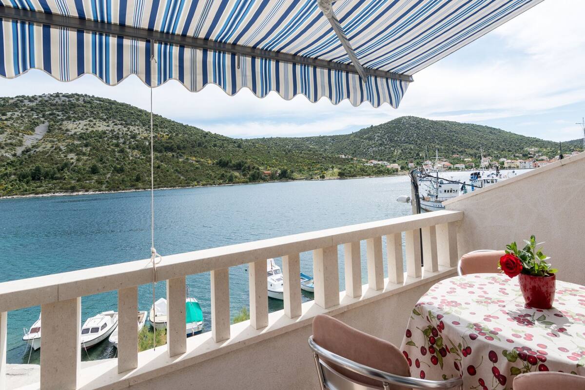 Un balcon doté d’un auvent rayé, d’où l’on peut admirer un plan d’eau, des bateaux et un paysage vallonné.
