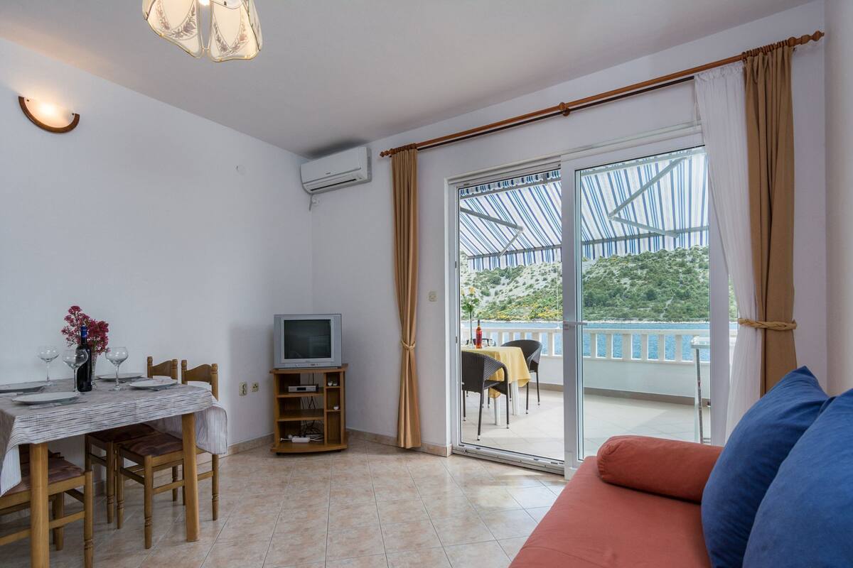 Une pièce comprenant une table à manger dressée pour deux personnes, un téléviseur posé sur un meuble, un canapé avec des coussins et une porte-fenêtre coulissante donnant sur un balcon extérieur avec vue sur la verdure et l’eau.