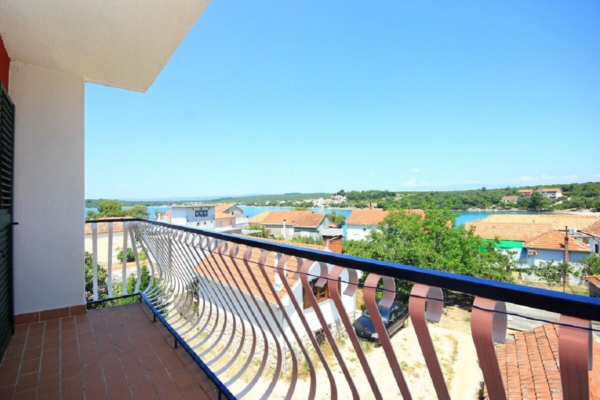 Ein Balkon mit geschwungener Metallreling, der auf einen Wohnbereich mit Häusern und einem Gewässer blickt.