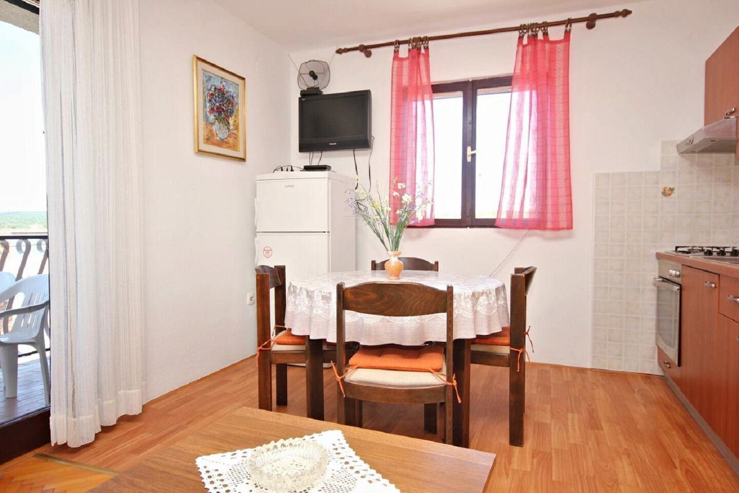 A dining area with a table set for four, a white refrigerator, a television above the table, and a window with pink curtains.