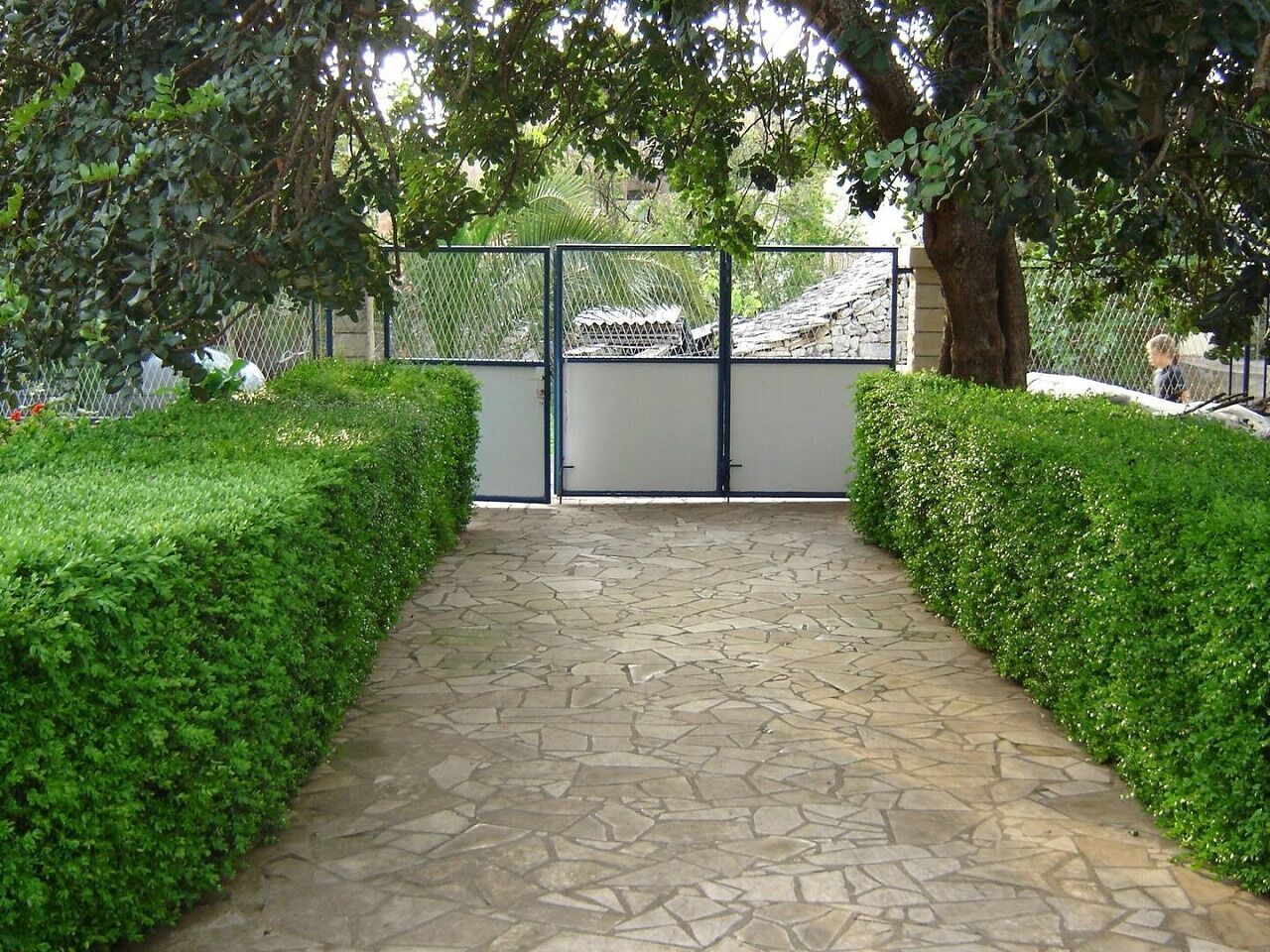 A stone pathway leading to a gated entrance, flanked by neatly trimmed hedges.
