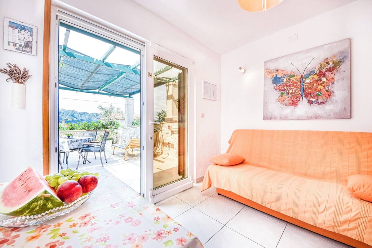 A living room with a sofa, a table with fruit, a glass door leading outside, and a butterfly painting on the wall.