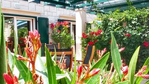 Un'area giardino con piante in vaso e fiori, una panchina in legno e l'ingresso di un edificio con tenda da sole verde.