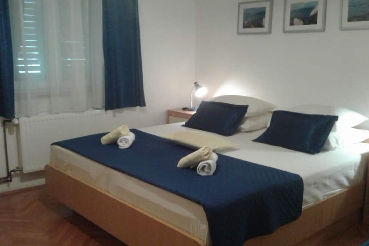 A double bed with a blue bedspread and two rolled towels, a bedside lamp, and a nightstand with a lamp.
