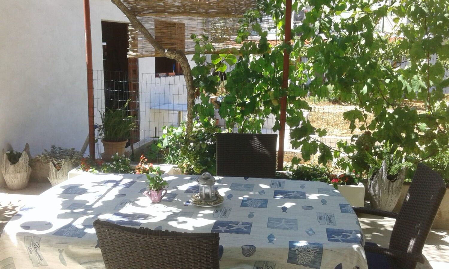 Une terrasse extérieure dotée d’une table ronde recouverte d’une nappe à motifs nautiques, entourée de chaises en osier.