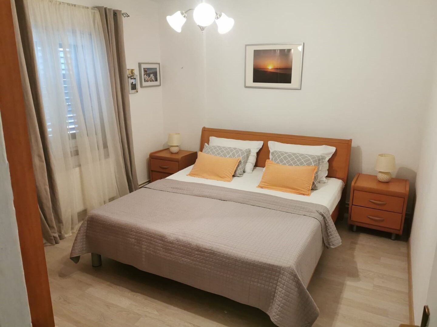 A neatly arranged bedroom with a bed, two bedside tables, a nightstand, a wall-mounted picture, and a window with curtains.