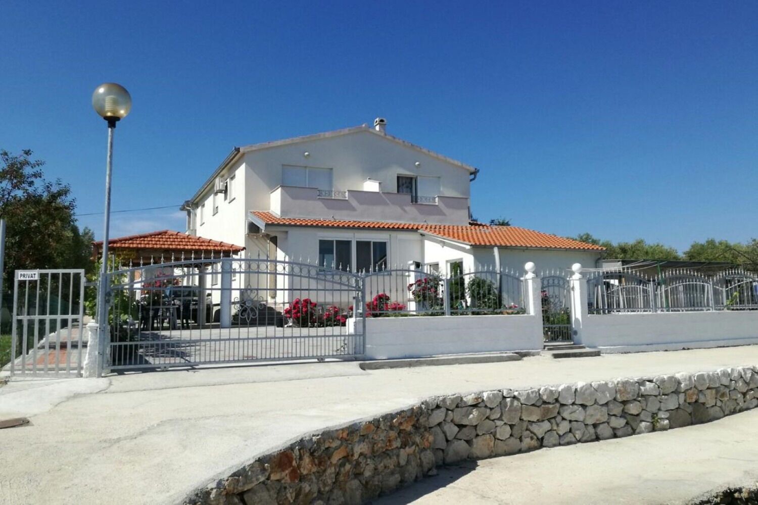 A two-story house with a white exterior and red-tiled roof, surrounded by a stone wall and metal gate.