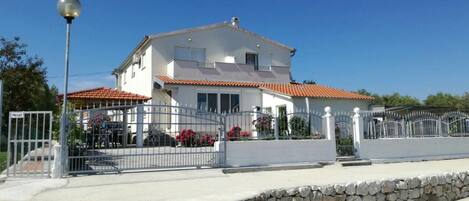 A two-story house with a white exterior and red-tiled roof, surrounded by a stone wall and metal gate.