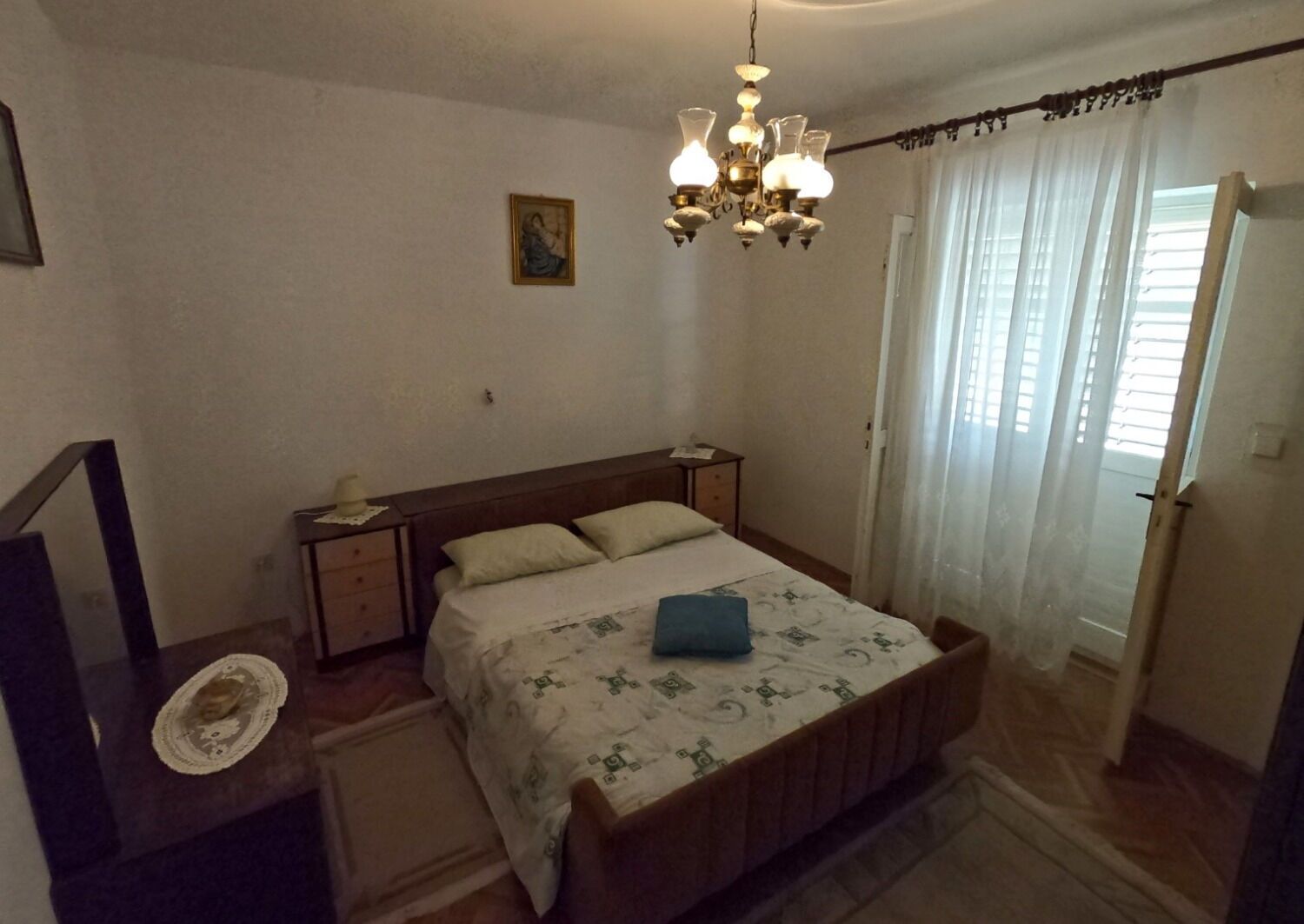 A bedroom with a wooden bed, a nightstand, a chair, a chandelier, and a window with curtains.