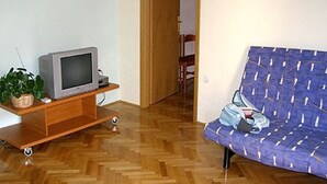 Una habitación con piso de madera, un sofá, un televisor sobre un mueble de madera y una puerta que da a otra habitación.