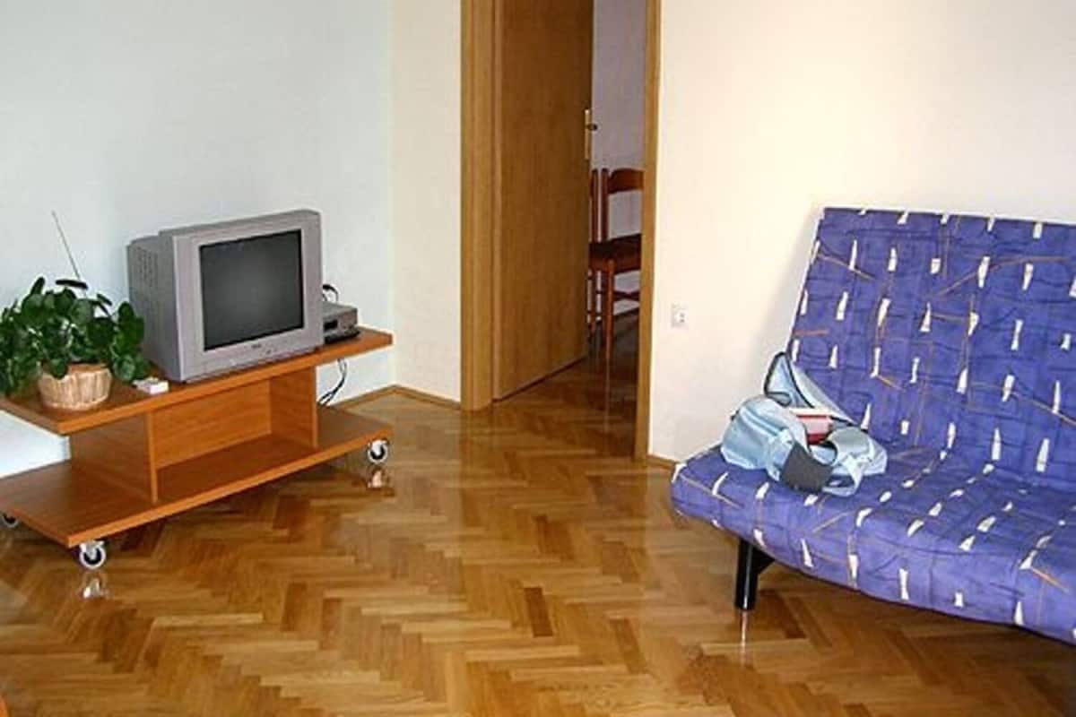 Una habitación con piso de madera, un sofá, un televisor sobre un mueble de madera y una puerta que da a otra habitación.