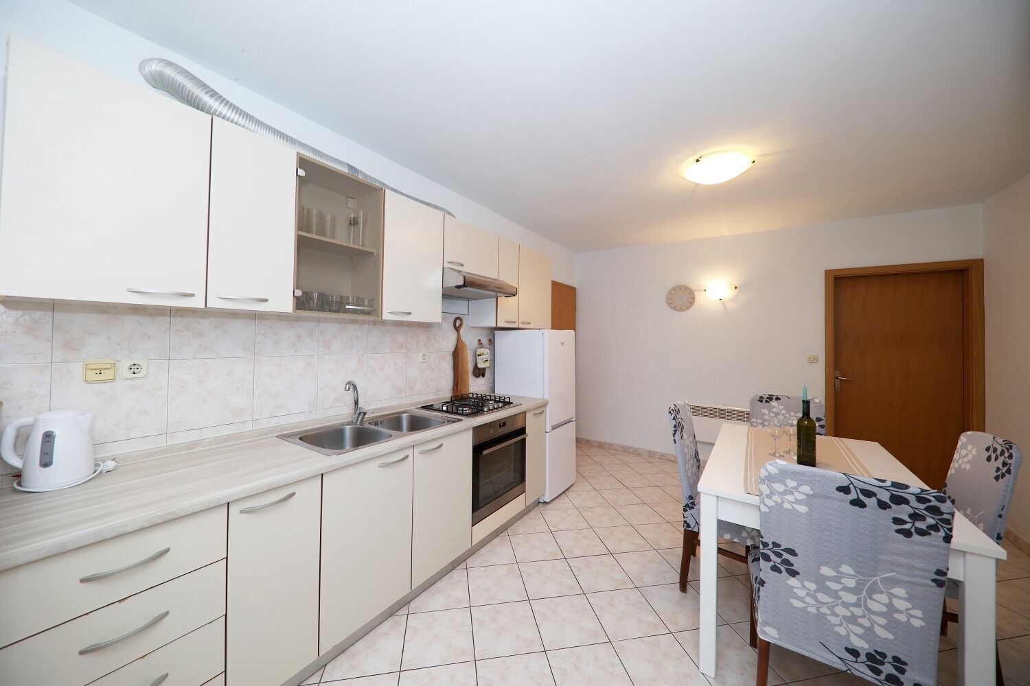 A compact kitchen with white cabinets, a gas stove, and a sink. There is a dining table with chairs and a small table lamp.