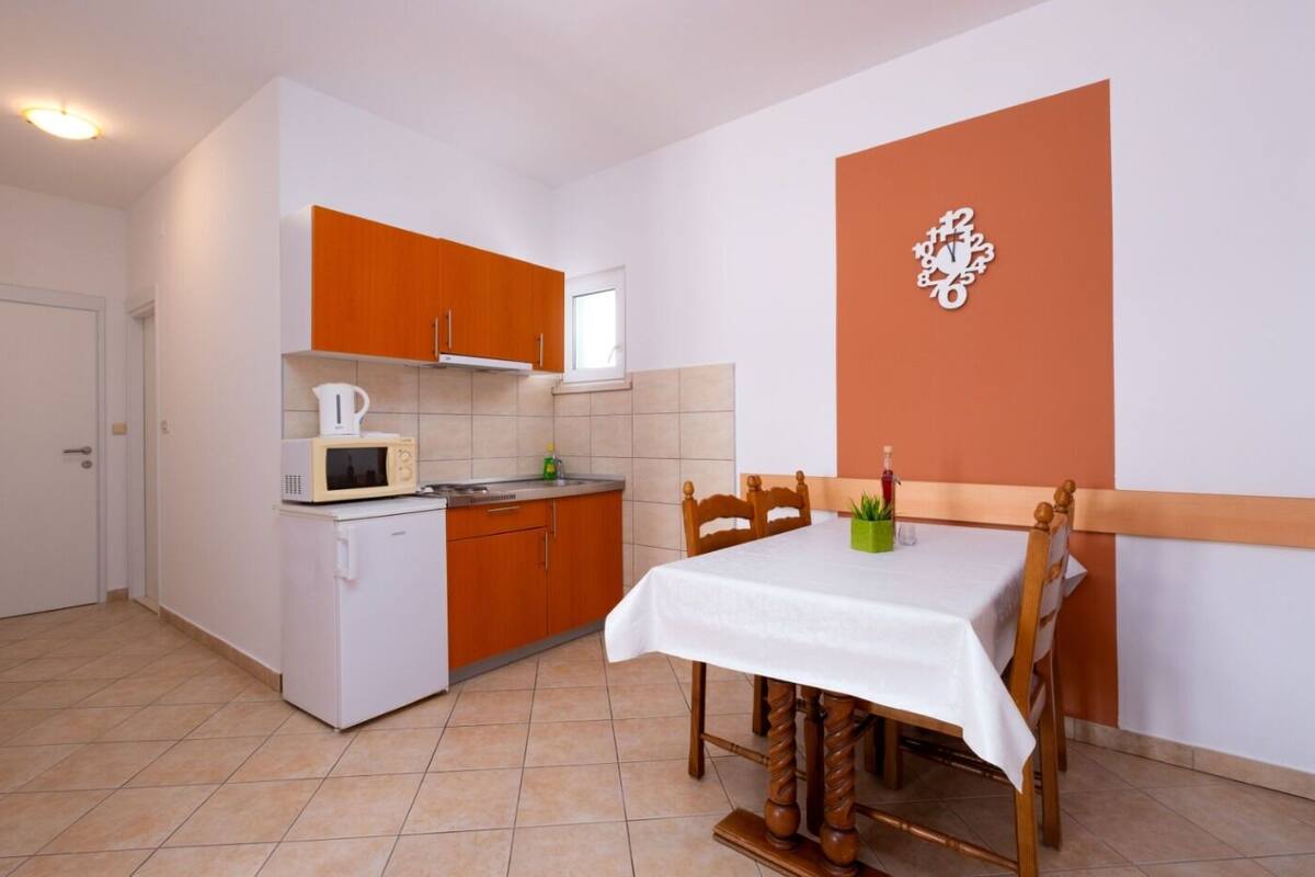 A compact kitchen with a microwave, kettle, and sink, a dining table with chairs, and a clock on the wall.