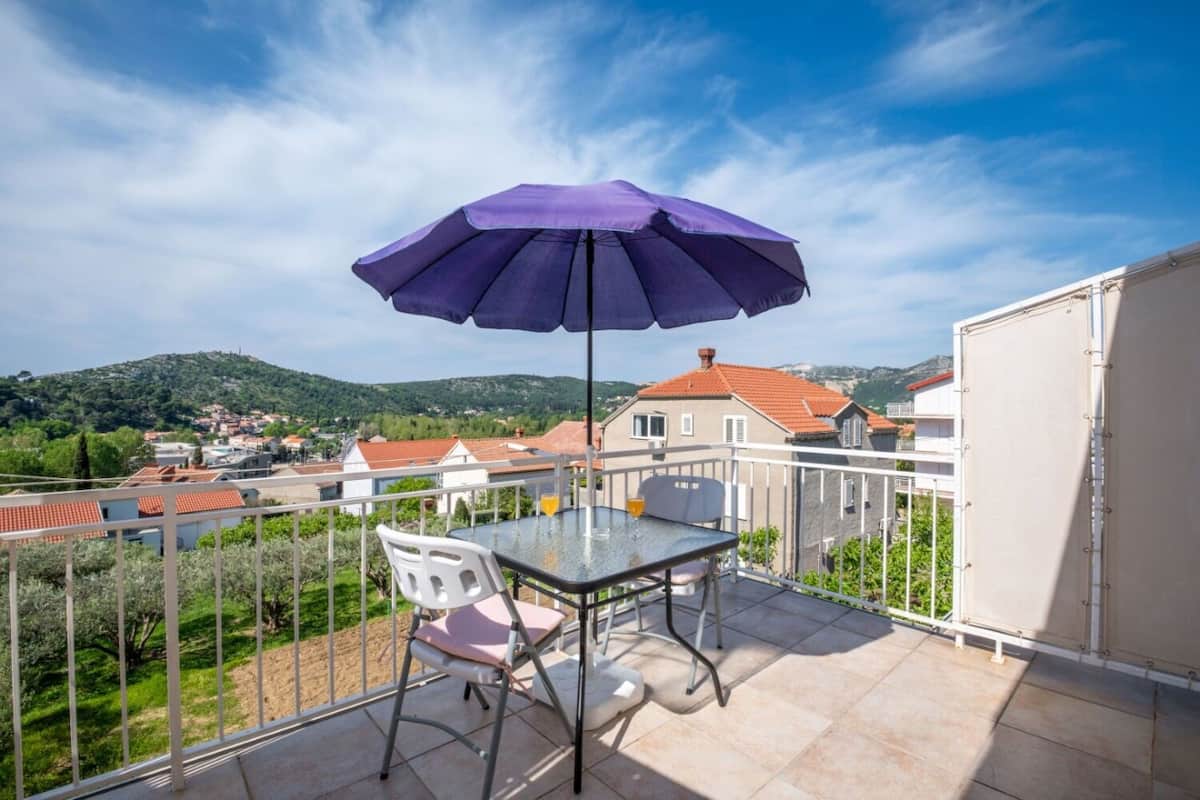 A balcony with a table and chairs, a purple umbrella, and a view of a town and hills.