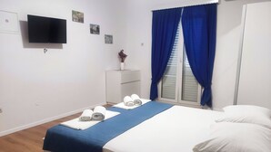 A neatly made bed with white linens and a blue blanket, a flat-screen TV mounted on the wall, a small bedside table with a vase, and a window with blue curtains. - Apartments Mat (Zupa dubrovacka)
