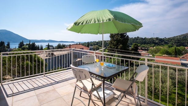 A balcony with a table and chairs, a green umbrella, and a view of a lake and mountains. - Apartments Mat (Zupa dubrovacka)