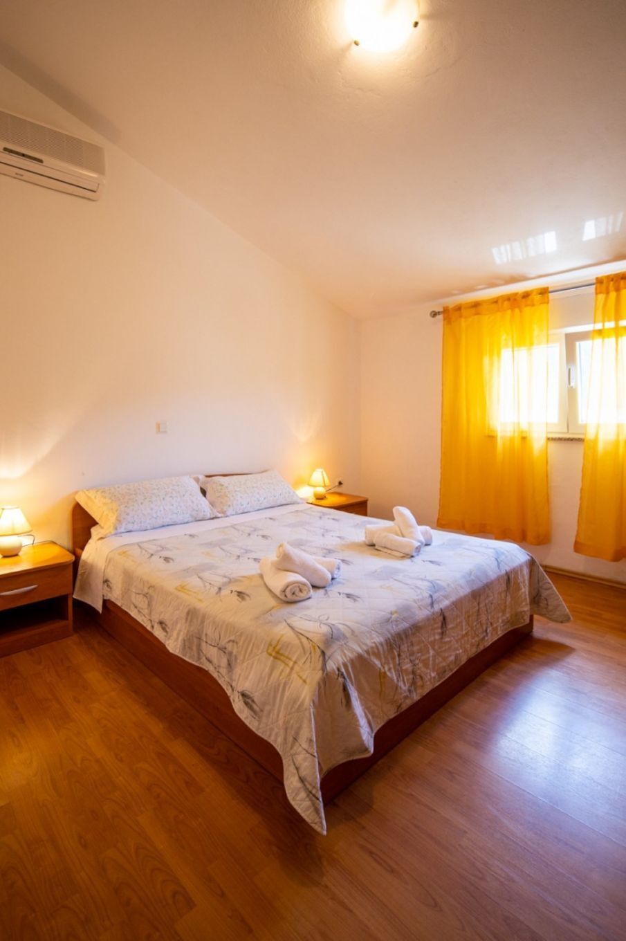 A bedroom with a wooden floor, a bed with a floral bedspread, two bedside tables with lamps, and a window with yellow curtains.
