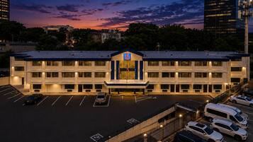 A multi-story hotel with a parking lot, a prominent entrance with a blue sign, and a sunset sky in the background.