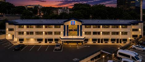 Un hotel de varios pisos con estacionamiento, una entrada destacada con un letrero azul y un cielo al atardecer de fondo.