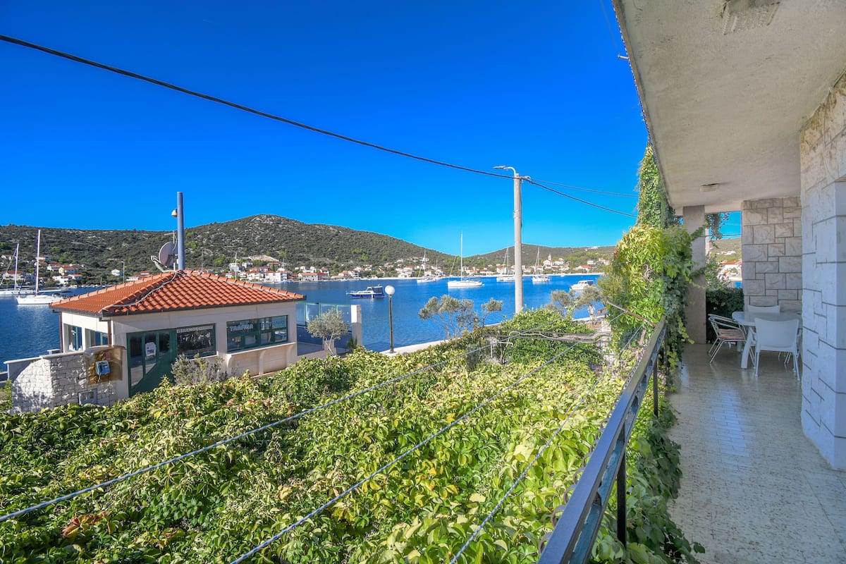 Un balcon donnant sur un port, un bâtiment avec un toit recouvert de tuiles rouges et un paysage montagneux en arrière-plan.