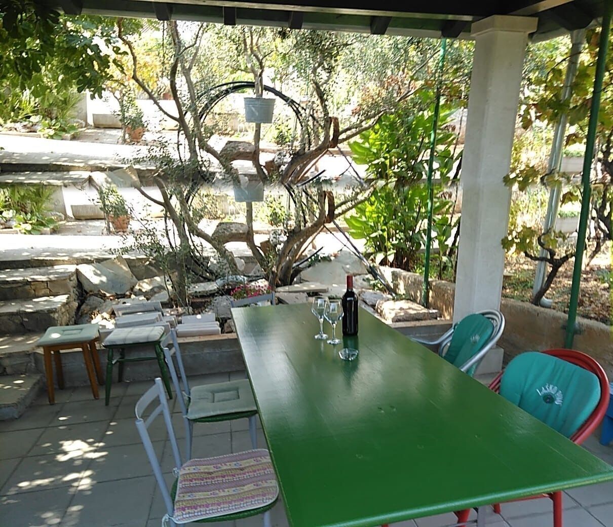 Una veranda esterna con un tavolo verde, sedie e una bottiglia di vino.